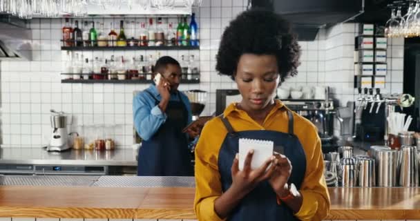 Afro-americano joven camarera bonita de pie en la cafetería y escribir lista de pedidos o menú de anotación. Hombre camarero compañero de trabajo hablando en el teléfono móvil en segundo plano. En el trabajo. — Vídeos de Stock