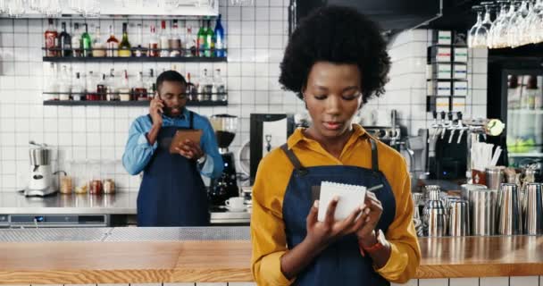 Portrét veselé mladé krásné ženy, usmívající se do kamery. Afroameričanka hezká servírka stojící v kavárně a na psacím listu nebo v menu. Muž kolega číšník mluví na mobilu na pozadí — Stock video