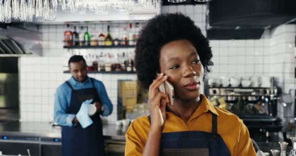 Bela jovem garçonete afro-americana falando no celular no balcão no bar. Mulher barista a falar ao telemóvel. Garçom limpando óculos no fundo. Copos de limpeza em café. — Vídeo de Stock