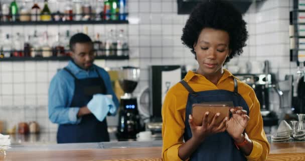 Csinos, fiatal afro-amerikai pincérnő, aki a bárpultnál telefonál. Női barista sms-t küld az okostelefonon. A pincér poharakat törölget a háttérben. Kelyhek tisztítása a kávézóban — Stock videók