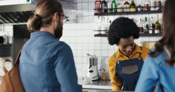 カフェでコーヒーを注文し、アフリカ系アメリカ人女性のバリスタと話す客の白人カップルの後部。顧客、男性と女性のためのカウンターでサービスを待っています。混合レース。多民族. — ストック動画