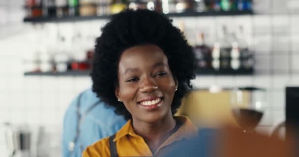 アフリカ系アメリカ人のウエイトレスの笑顔を閉じ、カフェでカウンターでクライアントと話しています。お客様とお話ができた幸せな陽気な女性バリスタ。女バーテンダーが笑っておしゃべり. — ストック動画