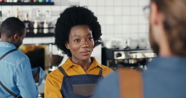 Afrikansk amerikansk servitris ler och pratar med en vit man vid disken på kaféet. Kvinna barista har trevligt prata med kunden. Kvinnlig bartender chattar med en manlig klient. Blandraser. Multietniskt — Stockvideo