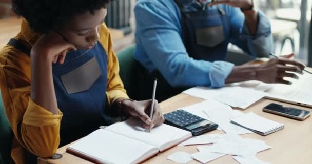 アフリカ系アメリカ人のカップル、バーテンダーが机に座ってノートブックのビジネス計算書を書いています。男性と女性はカフェの支出と損害を計算します。ロックダウン後に再起動。数え上げリストを作る. — ストック動画