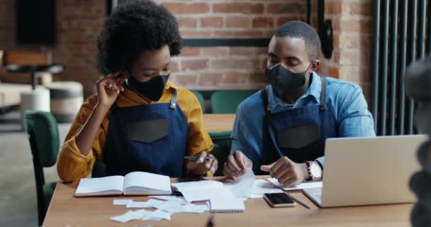 Maskeli Afro-Amerikalı çift restoran hesaplamaları yapıyor ve faturaları kontrol ediyor. Harcamalar ve hasarlar hesaplanıyor. Tecritten sonra tekrar açın. Dizüstü bilgisayarda sayılıyor Küçük girişimcilik — Stok video