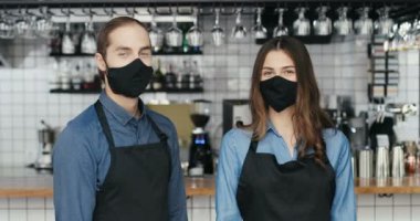 Maskeli ve eldivenli genç beyaz barmenlerin birbirlerine bakıp kameraya mutlu bir şekilde gülümsedikleri portre. Kafedeki mutlu garson ve garson. Neşeli erkek ve kadın, iş yerinde baristalar..