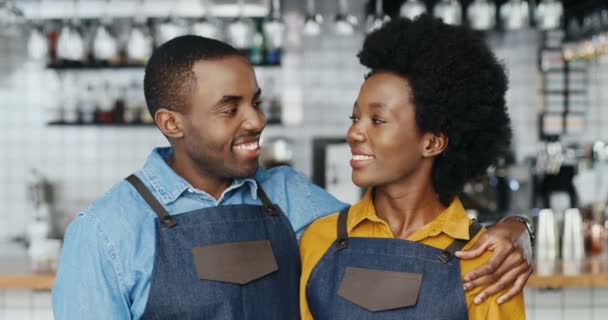 Retrato de un alegre par de camareros afroamericanos en delantales abrazándose y sonriendo a la cámara felizmente. Feliz camarero y camarera en abrazos en la cafetería. Hombre alegre abrazando a la mujer. Baristas. — Vídeos de Stock