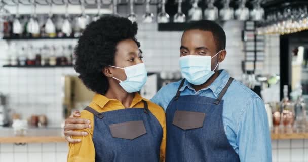 Retrato de un alegre par de camareros afroamericanos con máscaras médicas abrazándose, sonriendo a la cámara felizmente. Feliz camarero y camarera en abrazos en la cafetería. Hombre alegre abrazando mujer baristas — Vídeos de Stock