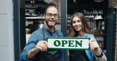 Önlüklü, kameraya mutlu bir şekilde gülümseyen ve kafenin girişinde masaları açık tutan beyaz barmenlerin portresi. Bar kapısında panosu açık, mutlu garson ve garson. Neşeli erkek kadın..