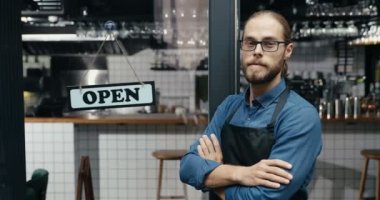 Kafe girişindeki masası açık kameraya gülümseyen önlüklü ve gözlüklü yakışıklı, mutlu, genç bir garson portresi. Neşeli, gülümseyen, barın kapısında tahtası açık duran erkek barmen.