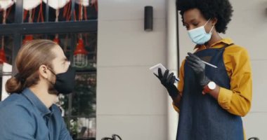 Kafeteryadaki beyaz erkek müşteri ve maskeli Afro-Amerikan bayan barista konuşuyor. Garson, açık hava restoranındaki müşteriden sipariş alıyor. Mi-x-ırkları. Çok ırklı. Salgın.