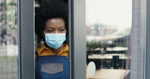 Afrikai-amerikai fiatal csinos pincérnő kinyitja a kávézó ajtaját, és kijön az utcára friss levegőt szívni. Közelkép egy gyönyörű női baristáról, aki kimegy. Újranyitás a világjárvány idején. — Stock videók
