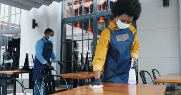 Pareja afroamericana de baristas, macho y hembra en máscaras limpiando escritorios en cafetería con spray desinfectante. Camarero y camarera desinfectando mesas en la cafetería. Desinfección. Vuelva a abrir en el bloqueo. — Vídeo de stock