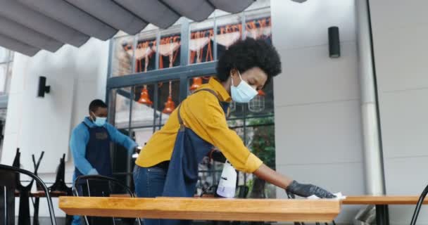 Casal afro-americano de baristas em máscaras de limpeza de mesas em bar com higienizadores spray. Garçom e garçonete desinfectando mesas no café. Desinfecção do local público. Reabrir após coronavírus. — Vídeo de Stock