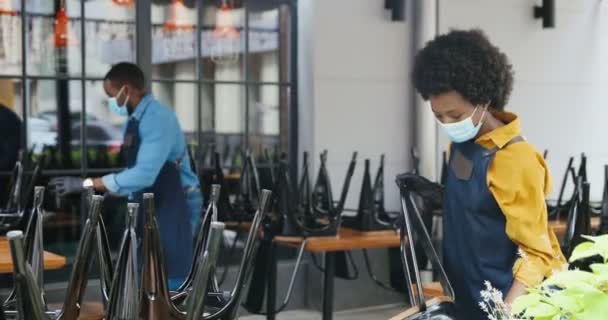 Pareja afroamericana de baristas, hombre y mujer en máscaras y guantes limpiando escritorios y sillas en el bar. Camarero y camarera limpiar y preparar lugar público en la cafetería. Concepto de reapertura. Preparación. — Vídeo de stock