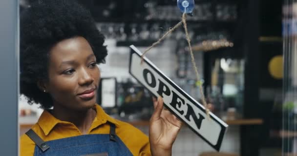 Közelkép afro-amerikai gyönyörű boldog nő pincér változó asztal az ajtón - zárva a kávézóban reggel. Női nyitó bár korán és mosolyogva. Szép eladó esztergálás fórumon, és megnyitja a boltot. — Stock videók