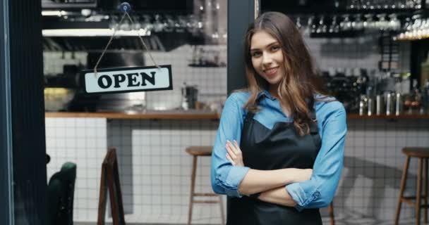 Portrait of beautiful Caucasian young happy woman waitress in apron smiling to camera with table open at cafe entrance outdoor. Pretty cheerful barista standing at bar door with board open. — Stock Video