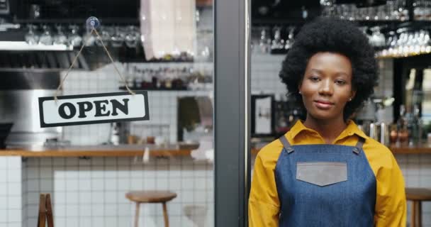 Portrait de la belle jeune serveuse afro-américaine heureuse en tablier souriant à la caméra avec table ouverte à l'entrée du café en plein air. Jolie barista joyeux debout à la porte du bar avec planche ouverte. — Video