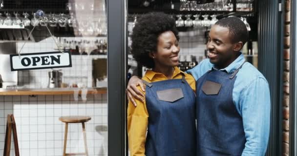 Porträt eines afroamerikanischen fröhlichen Barkeeperpaares in Schürzen, das sich umarmt und glücklich lächelt. Glücklicher Kellner und Kellnerin in Umarmungen an der Cafétür mit offenem Tisch. Fröhlicher Mann umarmt Frau. — Stockvideo