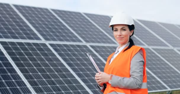 Retrato de joven ingeniera en uniforme especial de pie cerca de paneles solares. Hermosa profesional femenina en casco blanco sonriendo a la cámara. — Vídeos de Stock