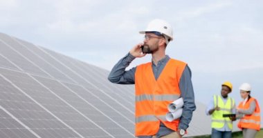 Yönetmen mühendisin portresi güneş çiftliğinde cep telefonuyla konuşuyor. İki işçi arka plandaki güneş panellerini inceliyor.