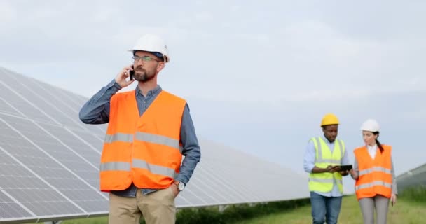 Retrato del ingeniero jefe hablando en un teléfono celular mientras estaba parado en la plantación solar. Dos trabajadores caminando mirando la tableta en el fondo. — Vídeos de Stock