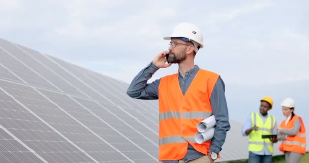 Retrato de engenheiro diretor falando no telefone celular em pé na plantação solar. Dois trabalhadores inspecionam painéis solares no fundo. — Vídeo de Stock
