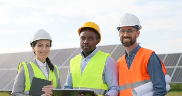 Portret inżynierów stojących na zewnątrz w pobliżu paneli słonecznych. Trzech pracowników: jedna kobieta i dwóch mężczyzn w specjalnym mundurze patrzą w kamerę i uśmiechają się. — Wideo stockowe