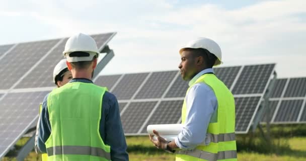 Tři dělníci ve speciální uniformě diskutují o pracovních chvílích venku. Jedna žena a dva muži mluví o solární plantáži. Týmová práce. — Stock video