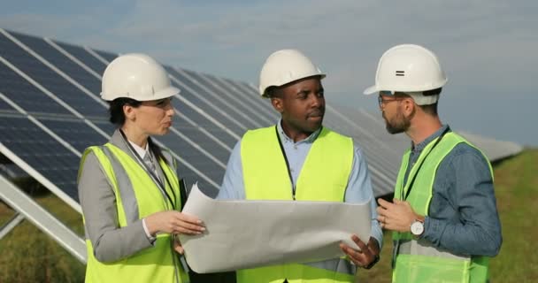 Tres ingenieros técnicos revisan el plan en papel y lo discuten haciendo gestos con las manos. Plantación solar. Equipo multinacional. — Vídeos de Stock