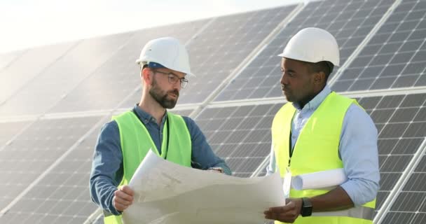 Dwóch inżynierów w mundurach dyskutuje o planie trzymania paneli słonecznych. Afro-amerykański technik słuchający wyjaśnień kierownika projektu białego człowieka. — Wideo stockowe