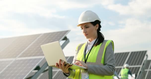 Portrait of beautiful female technologist in uniform with protective helmet.Adult woman holding laptop in her hands standing on field with solar panels. — Stock Video