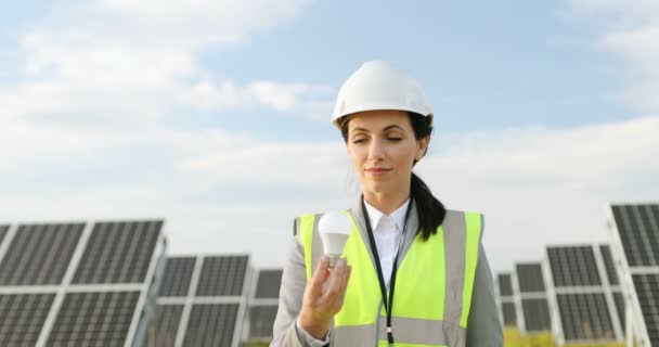 Portrait of female technologist in uniform with protective helmet among the field with solar panels. Beautiful woman reaches out showing light bulb. — Stock Video
