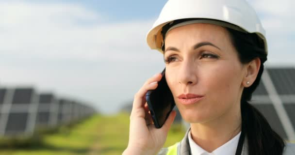 Close-up portret van mooie vrouwelijke technoloog praten op mobiele telefoon in beschermende witte helm staan op de buitenkant. Zonnepanelen op de achtergrond. — Stockvideo