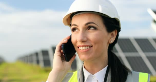 Close up portrait of beautiful female technologist talking on mobile phone among field of solar panels on background. — Stock Video