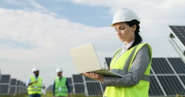 Bella donna d'affari in piedi tra il campo dei pannelli solari in uniforme speciale. Ingegnere femminile utilizza il computer portatile per lavorare all'esterno. — Video Stock