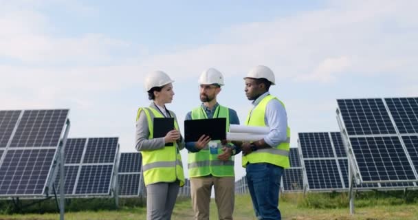 Três engenheiros em unifom: uma mulher e dois homens em pé entre painéis solares e discutir layout de instalações. — Vídeo de Stock