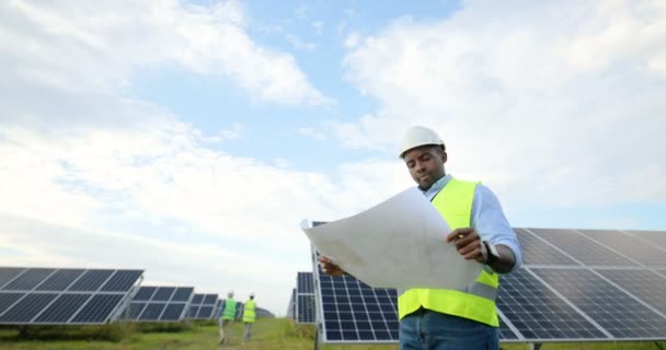 Портрет молодого афроамериканського інженера, який тримає сонячний план у своїх руках. Чоловік у спеціальній уніформі й захисний шолом вивчає паперові схеми.. — стокове відео