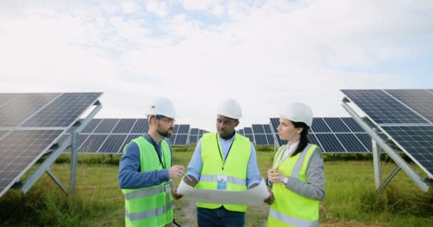 Tres ingenieros en uniformes especiales con cascos blancos discuten los procesos de trabajo en la granja solar. Dos hombres y una mujer están buscando un plan para la colocación de paneles solares. — Vídeos de Stock