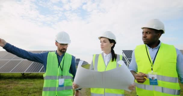 El hombre caucásico le dice a la mujer y al hombre afroamericano sobre trabajar en una granja solar. Mujer sosteniendo el plan de papel en sus manos. Tres ingenieros uniformados. — Vídeos de Stock