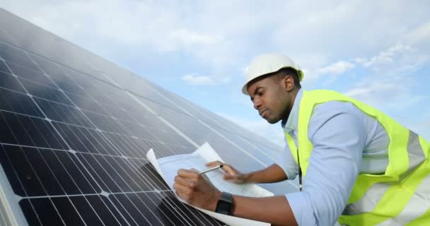 Ingeniero afroamericano en uniforme especial haciendo notas a lápiz en el diagrama de papel apoyado en el panel solar.Concepto de electricidad alternativa. — Vídeo de stock