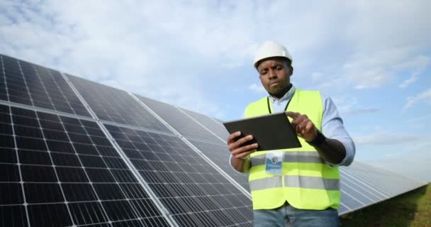 Афро-американский рабочий инженер в форме в белом шлеме идет рядами с солнечными панелями, делая заметки на планшете. — стоковое видео