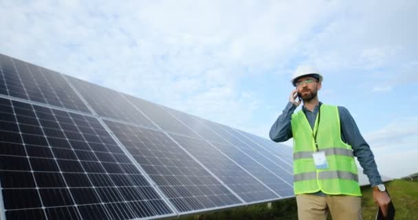 Porträt eines kaukasischen Ingenieurs beim Telefonieren. Solarfarmarbeiter läuft mit Batterien in der Hand durch die Reihen. — Stockvideo
