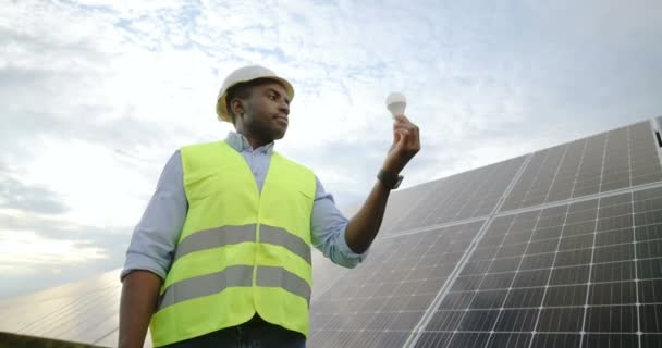 Afrikai amerikai mérnök portréja egyenruhában, védősisakkal, amint a villanykörtét nézi, amit a kezében tart. Az alternatív zöld energia fogalma. — Stock videók