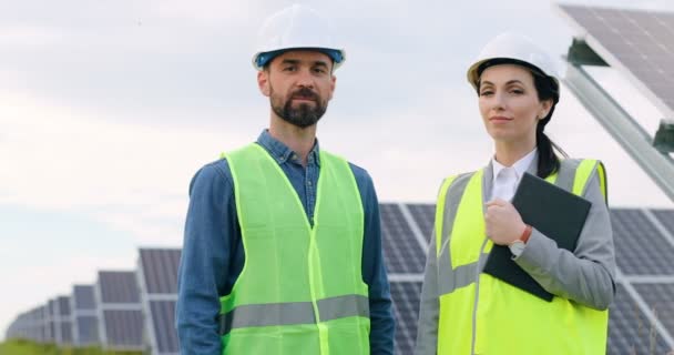 Portrét dvou inženýrů ve speciálních uniformách s ochrannými bílými přilbami. Krásná žena a běloch stojí mezi solárními panely a usmívá se. — Stock video