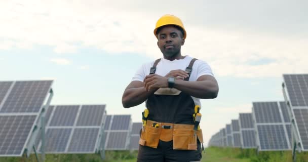 Retrato do jovem artesão afro-americano bonito em capacete de proteção. Homem de uniforme e com ferramentas em pé entre painéis solares. — Vídeo de Stock