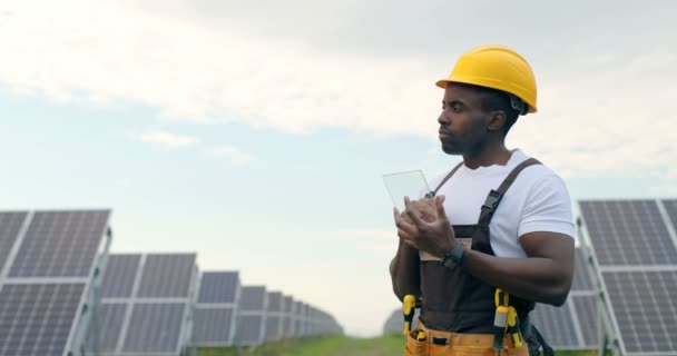 Portret młodego przystojnego Afroamerykanina mechanika w kasku ochronnym. Mężczyzna w mundurze trzyma tabliczkę w dłoniach i wprowadza tam dane.. — Wideo stockowe