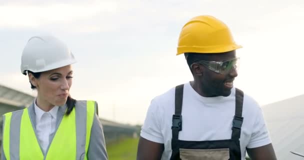 Three workers engineers in uniforms walk among rows of solar panels talking and joking. Concept of alternative electricity generation — Stock Video