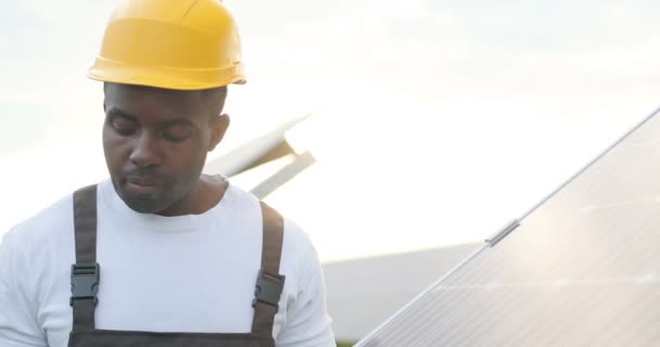 Close up of young African American mechanic in yellow helmet working drill on solar panel. Concept of green electricity. — Stock Video