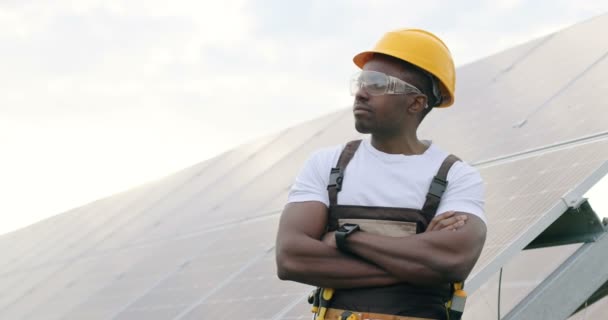 Handsome African-American mechanic in coveralls and protective yellow helmet standing outside cross-armed among solar panels and looks away. — Stock Video